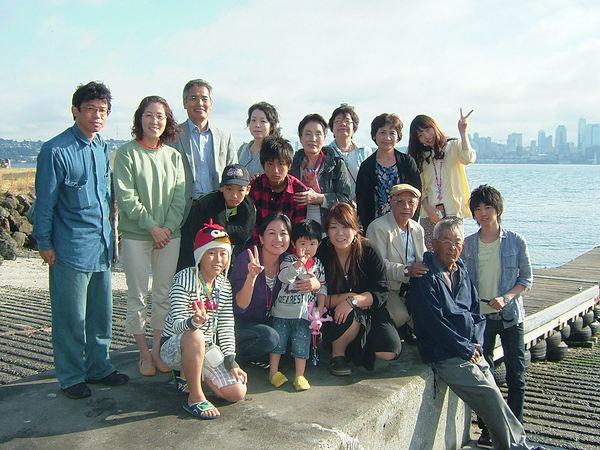 広い川の前で男性、女性、子供達17人みんなで一緒に集まりピースをしたり笑顔の集合写真